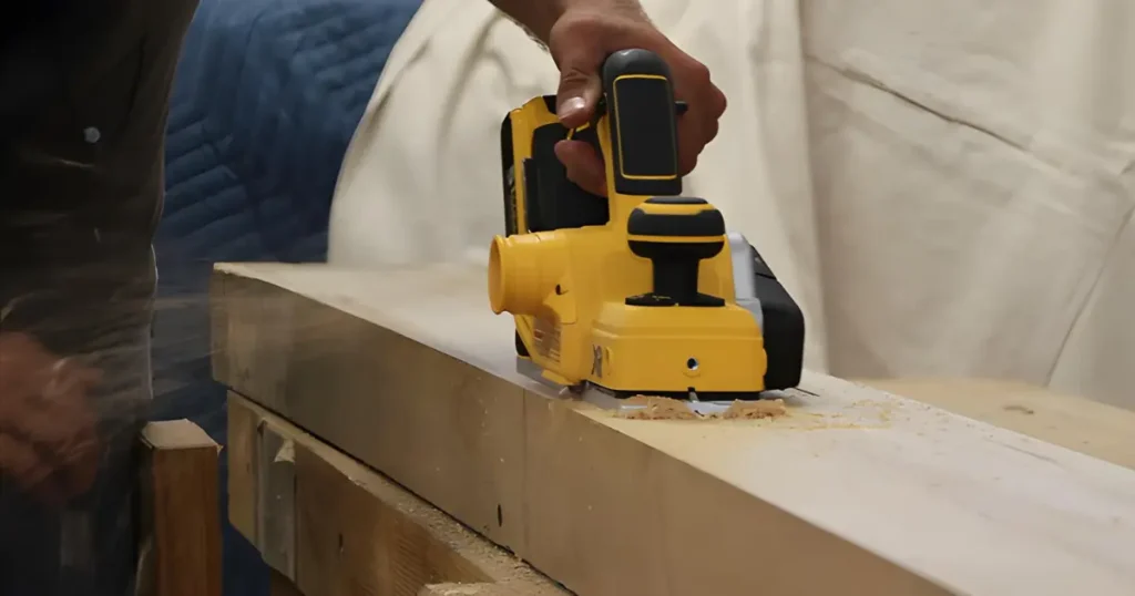 The DEWALT 20V MAX Cordless Planer (DCP580B) in use, guided by a woodworker to flatten the surface of a thick wooden beam, with chips ejecting.
