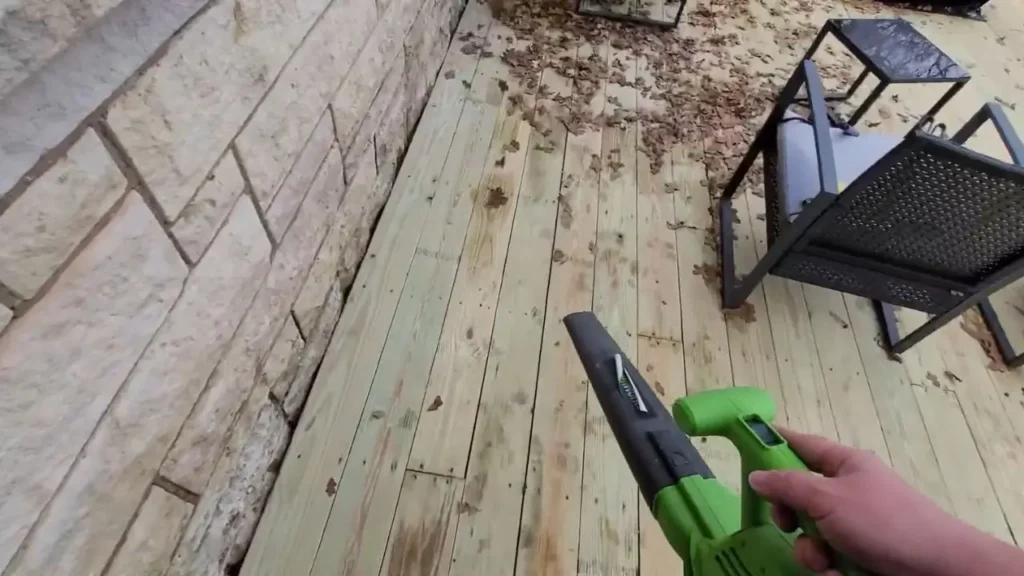 Person's hand holding Greenworks 24012 leaf blower, clearing leaves from a wooden deck, first-person view. Smart AI Gears test.
