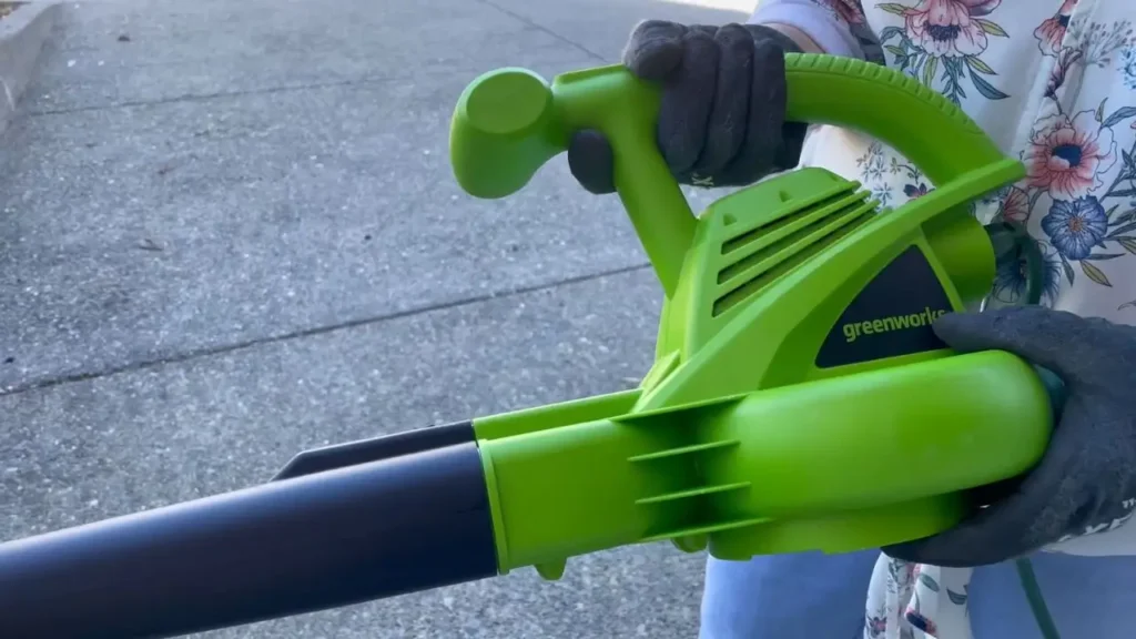 Hands wearing work gloves holding the Greenworks 24012 bright green electric leaf blower, showing comfortable grip. Smart AI Gears.