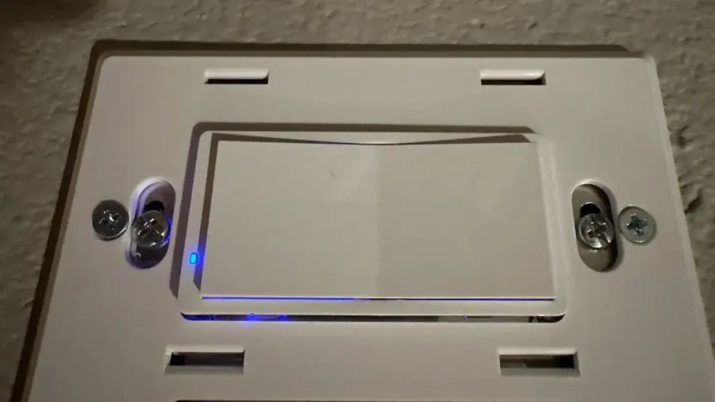 A close-up of a white Zooz Z-Wave Dimmer installed on a wall plate, with its small blue LED indicator light on.