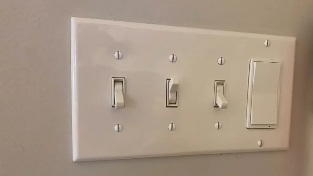 A white multi-gang wall plate showing three traditional toggle light switches next to one paddle-style smart dimmer.