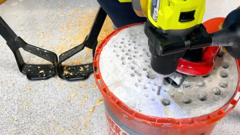 A drill with a long masonry drill bit is actively drilling into a white concrete surface inside a red bucket. Dust extraction tubing is visible, showing a messy but controlled work environment.