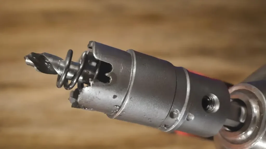 A close-up of a hole saw drill bit attached to a power drill, with visible spiral bit and spring mechanism.