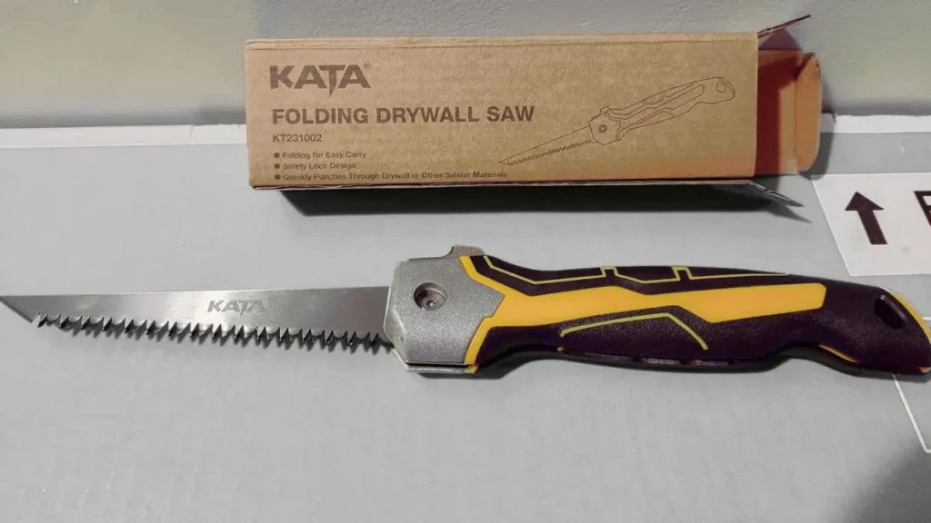 KATA Folding Jab Saw with a dual-tone yellow and black handle, safety lock system, and triple-ground teeth, placed beside its original box packaging.