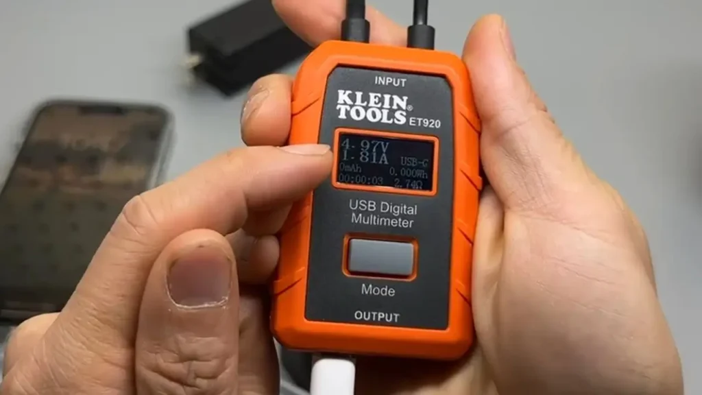Close-up of Klein Tools ET920 USB Meter display showing live voltage and current readings during a test, with a hand pointing at the screen.