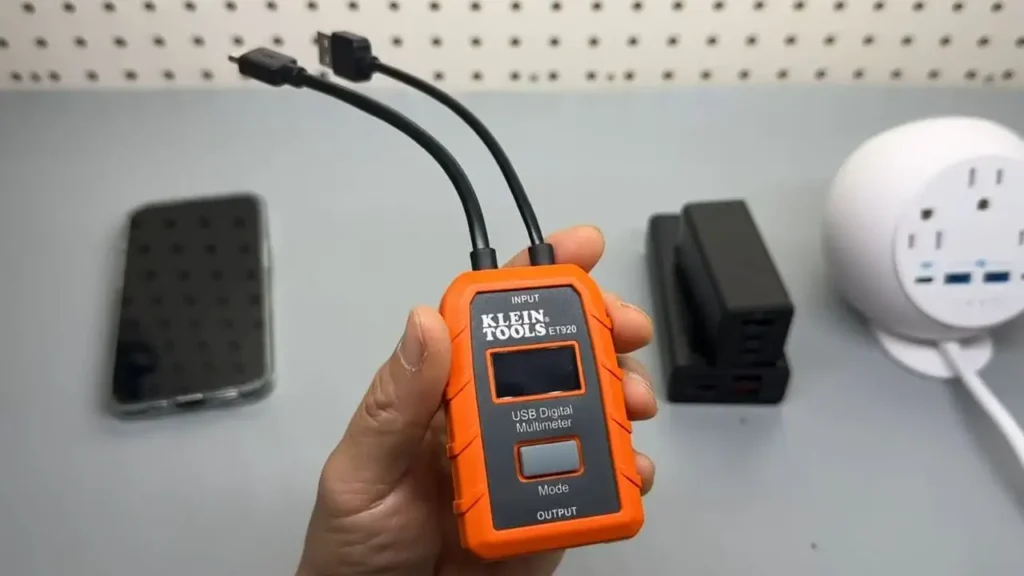 Klein Tools ET920 USB Meter review: Orange and black USB digital multimeter held in a hand with USB-A and USB-C cables, a smartphone, and chargers in the background, showing natural lighting and slight clutter. Smart AI Gears