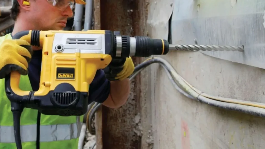 Construction worker drilling into a metal surface with a Dewalt hammer drill at a building site, wearing safety gear.