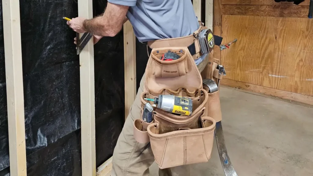 A carpenter wearing a leather framing tool belt pouch, which is filled with various tools, including a cordless drill and tape measure, while marking a wall stud with a pencil.