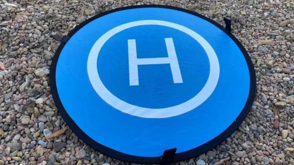 A blue and white KINBON landing pad is placed on a rough gravel surface.