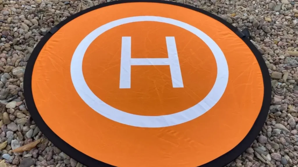 A bright orange and white KINBON landing pad is unfolded and placed on a gravel ground.