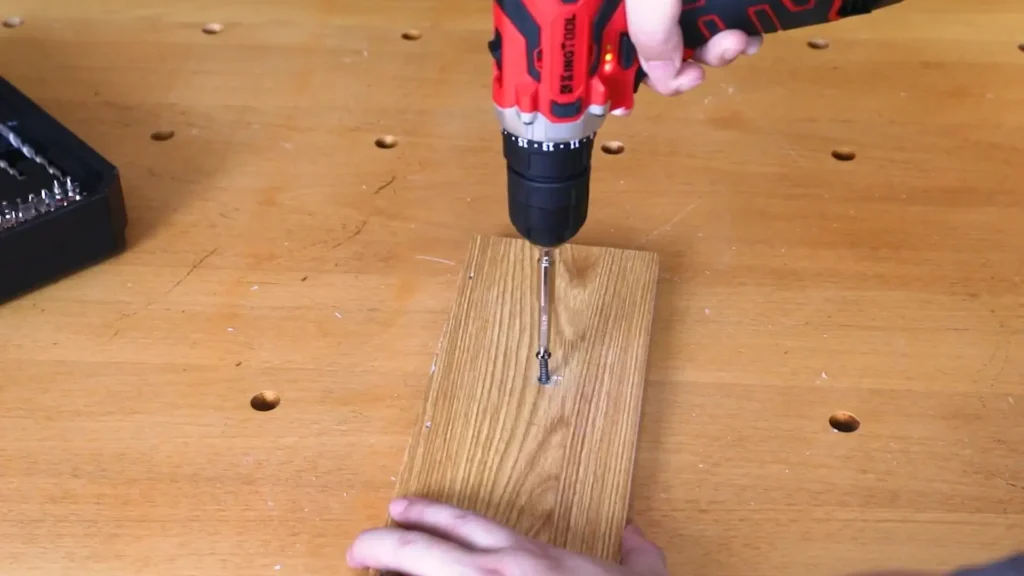 A top-down view of a person using a red cordless drill to drive a screw into a piece of light-colored wood on a workbench.