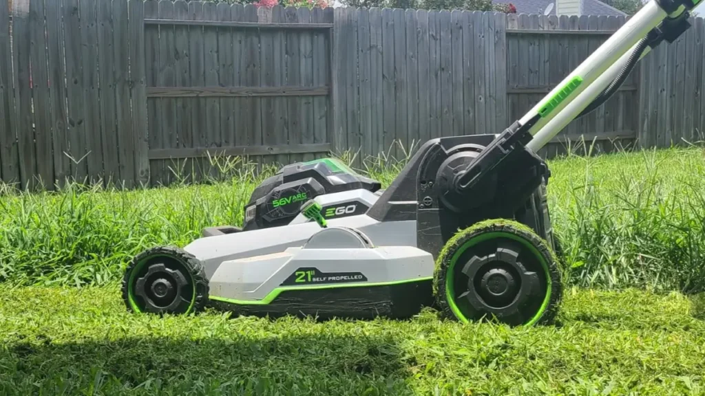A side view of a gray and green EGO 21-inch self-propelled mower cutting a clear path through tall green grass in a residential backyard.