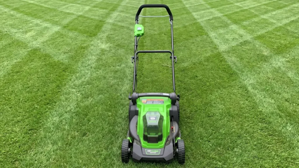 A front-facing view of a bright green and black Greenworks push lawn mower resting on a manicured grass lawn with visible diagonal mowing stripes.