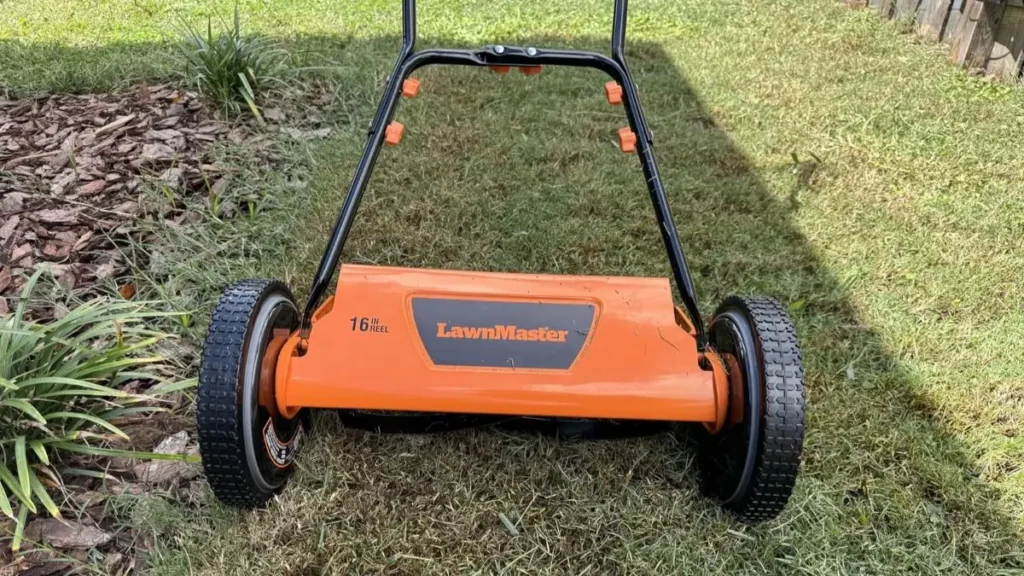 A front-facing view of an orange and black LawnMaster 16-inch manual push reel mower resting on a grassy lawn next to a mulched garden bed.