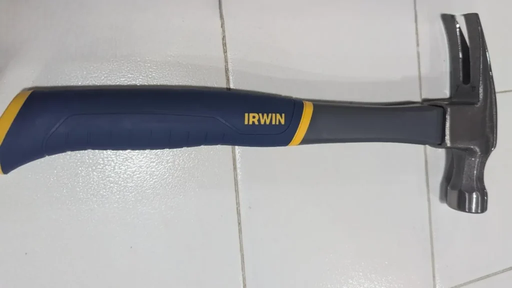 A close-up of the IRWIN 16oz fiberglass claw hammer laying on a white tiled surface, showing its steel head and blue and yellow rubberized handle.