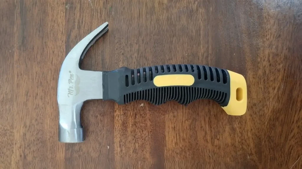 A close-up view of the Mr. Pen 8oz stubby claw hammer resting on a wooden surface, showcasing its short black and yellow rubber grip and steel head.